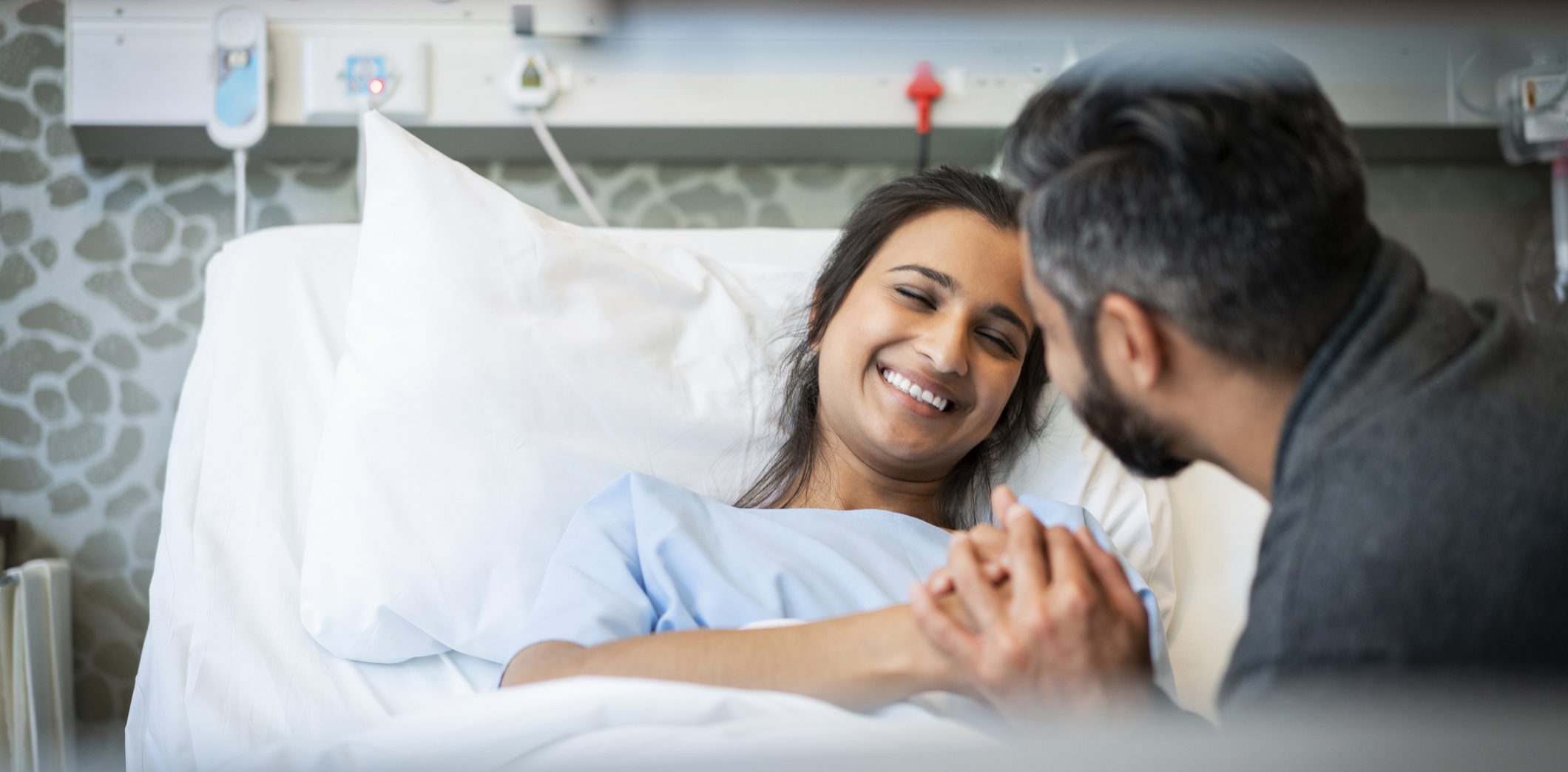 Man visiting smiling young woman in hospital ward. Loving male is holding her hand.