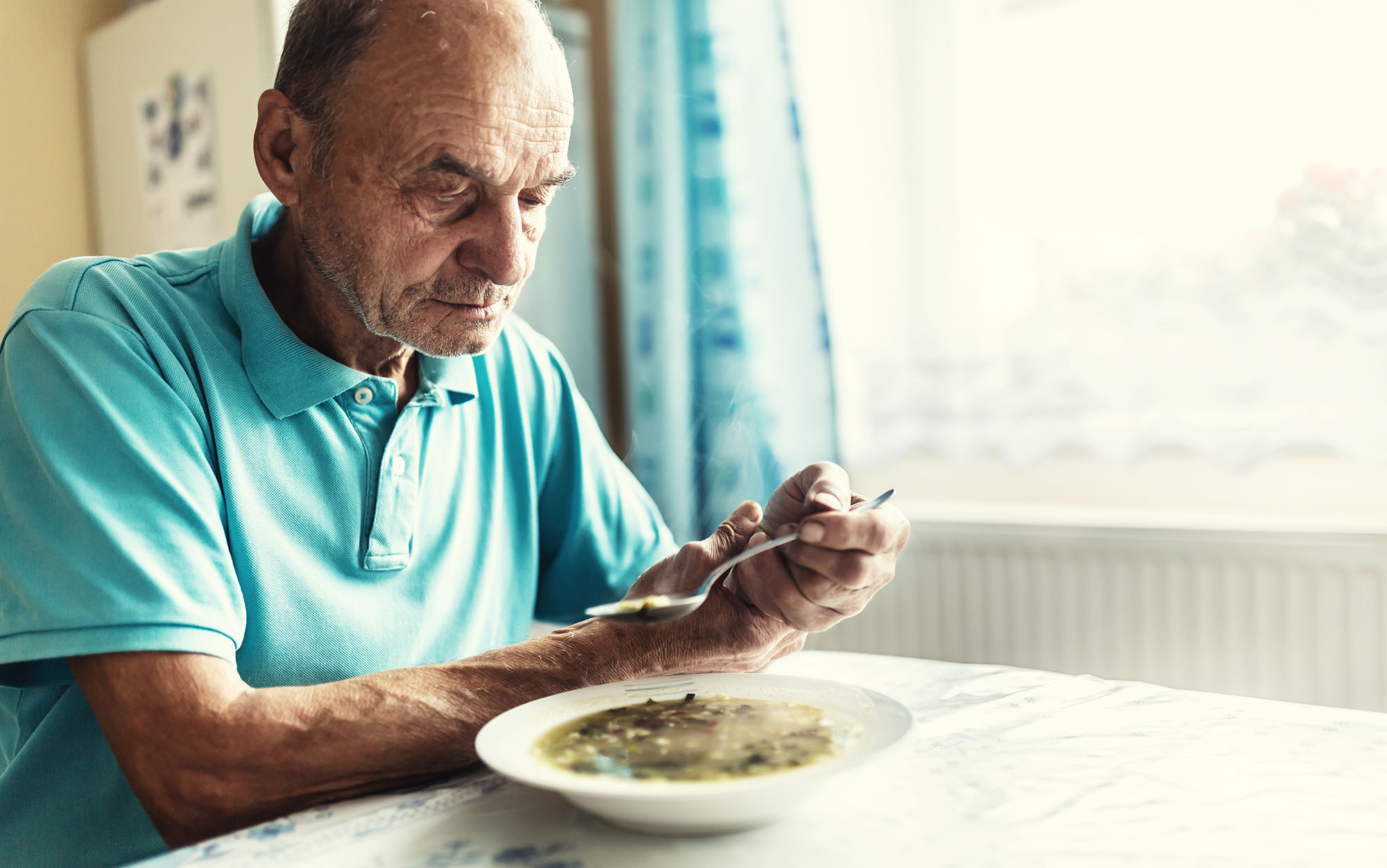 Senior man with a movement disorder or tremor holds his arm with a hand, trying to eat soup