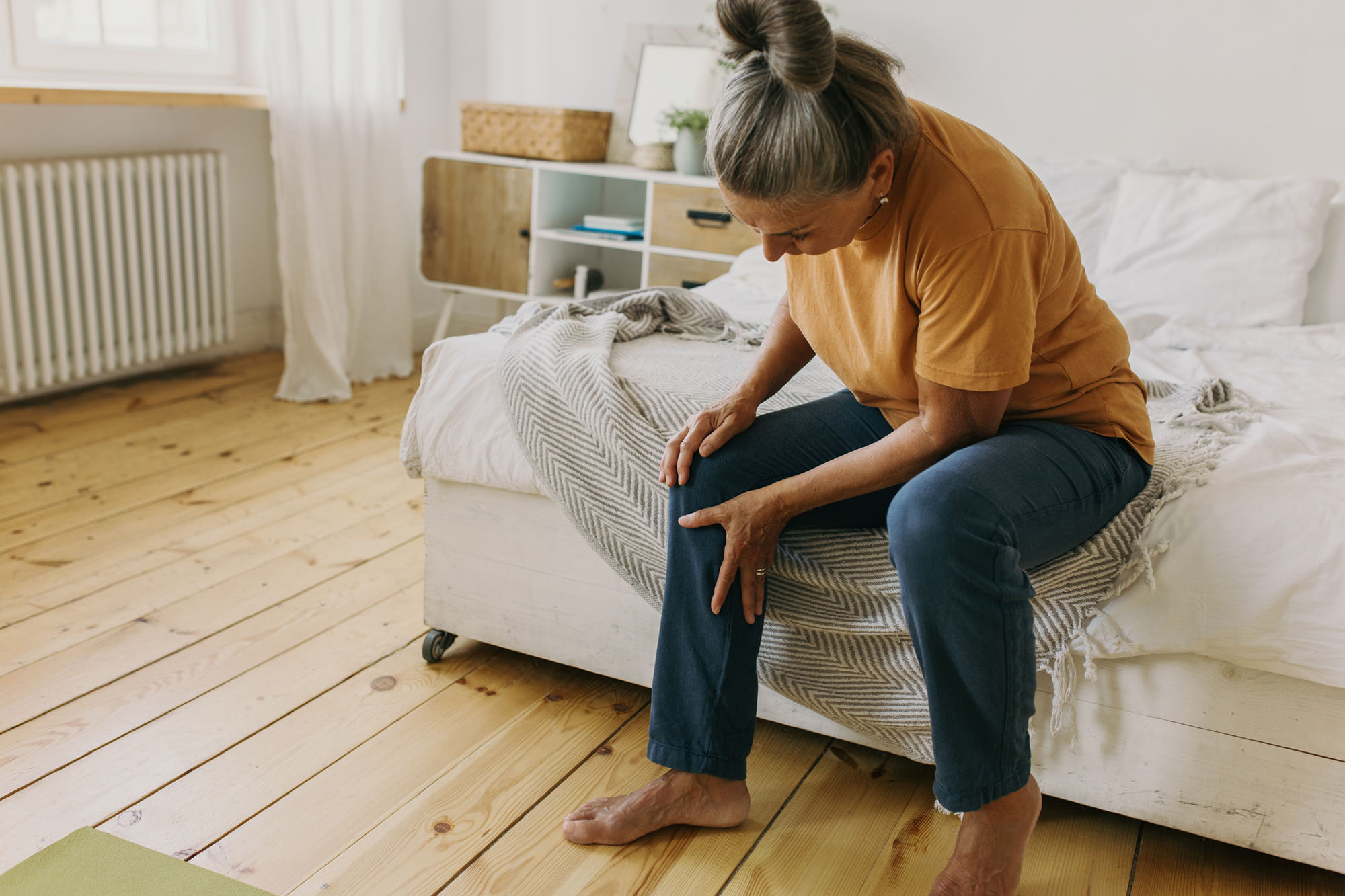 A senior woman sits on her bed and holds her right leg in some discomfort due to her knee.