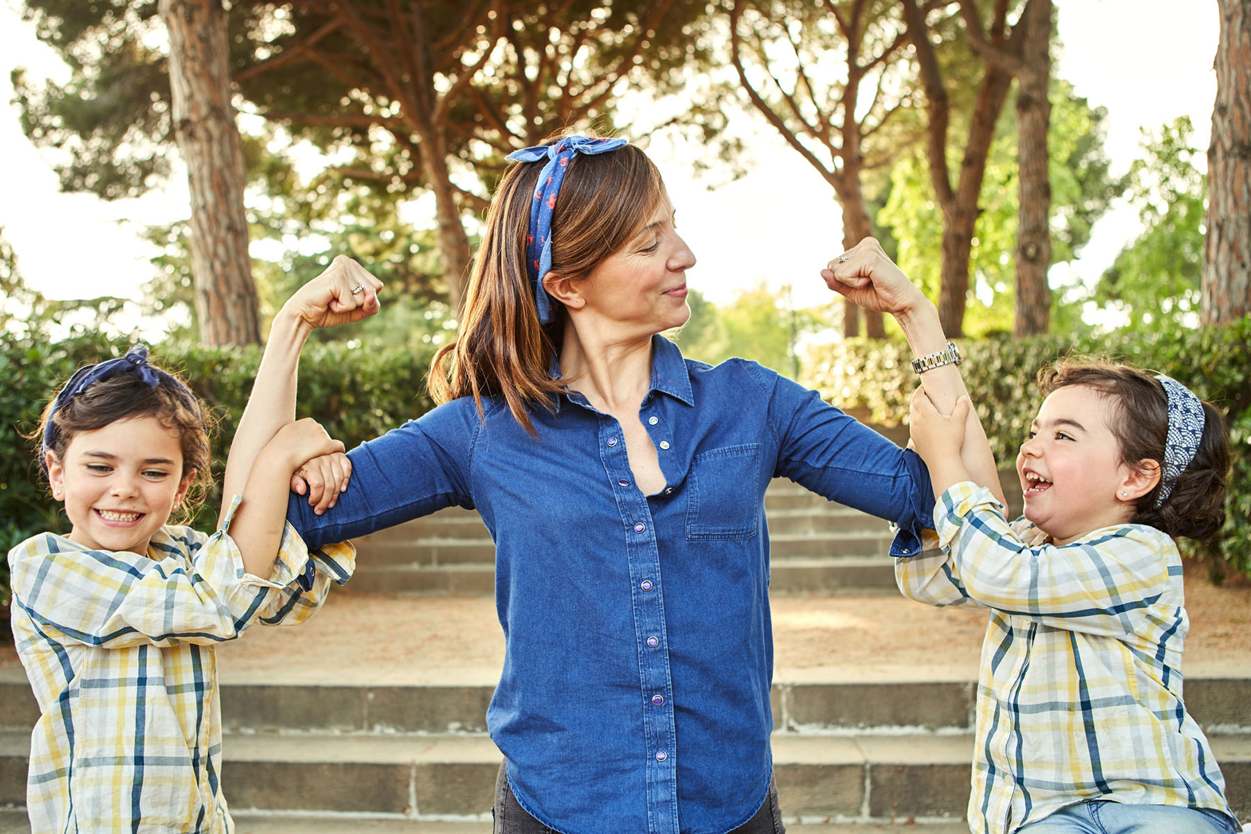 A young mom holds her arms up with her biceps curled as if to say girl power while her two young daughters hang onto each arm, all smiling