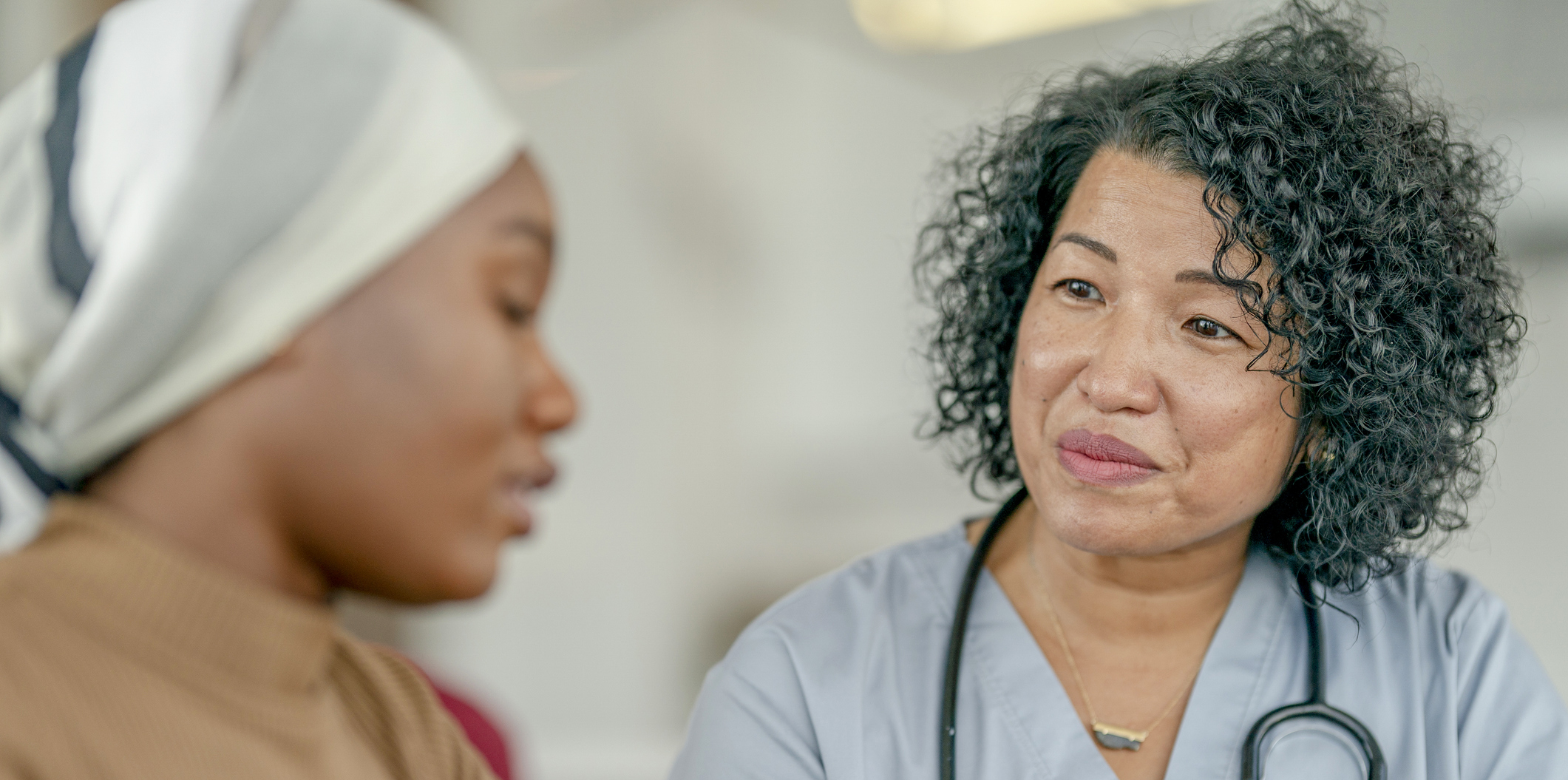 A female nurse of Asian decent, sits with her female cancer patient as she shares her recent test results with her. The patient is dressed casually and wearing a headscarf as she looks down somberly.