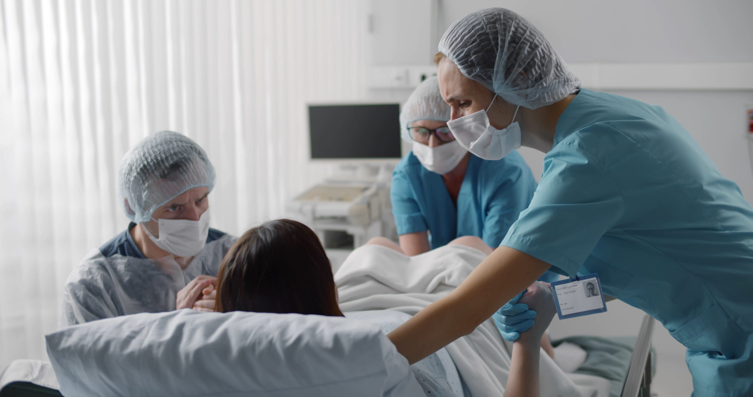 Woman giving birth with husband holds her hand in support and obstetricians assisting. Back view of medical staff in protective uniform helping pregnant woman in labor