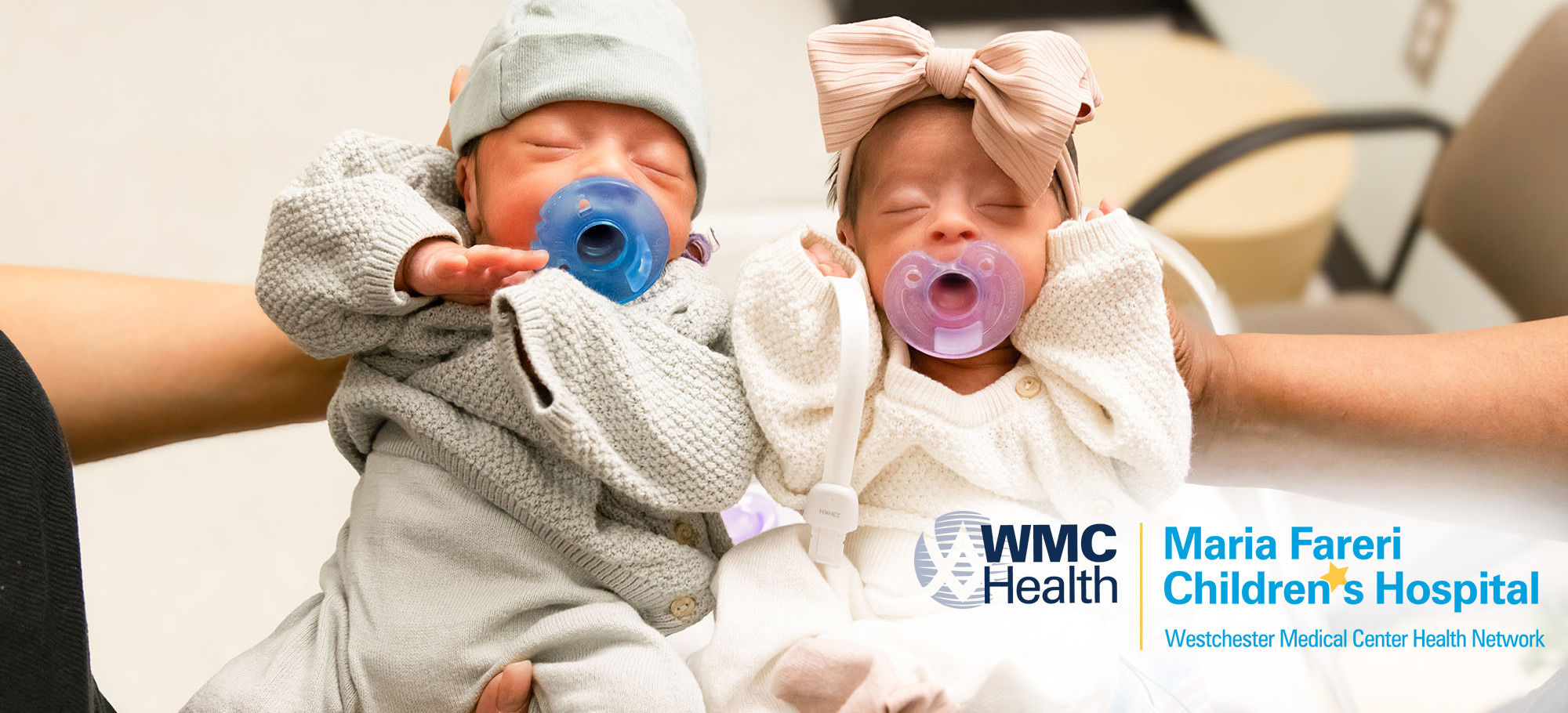 Two young newborn twin patients are held up by caregivers