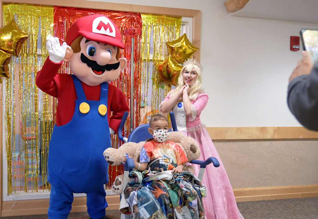 A children's hospital patient in a mask and on a wheelchair takes a photo with a person dressed as a Mario character and a person dressed as Princess Peach