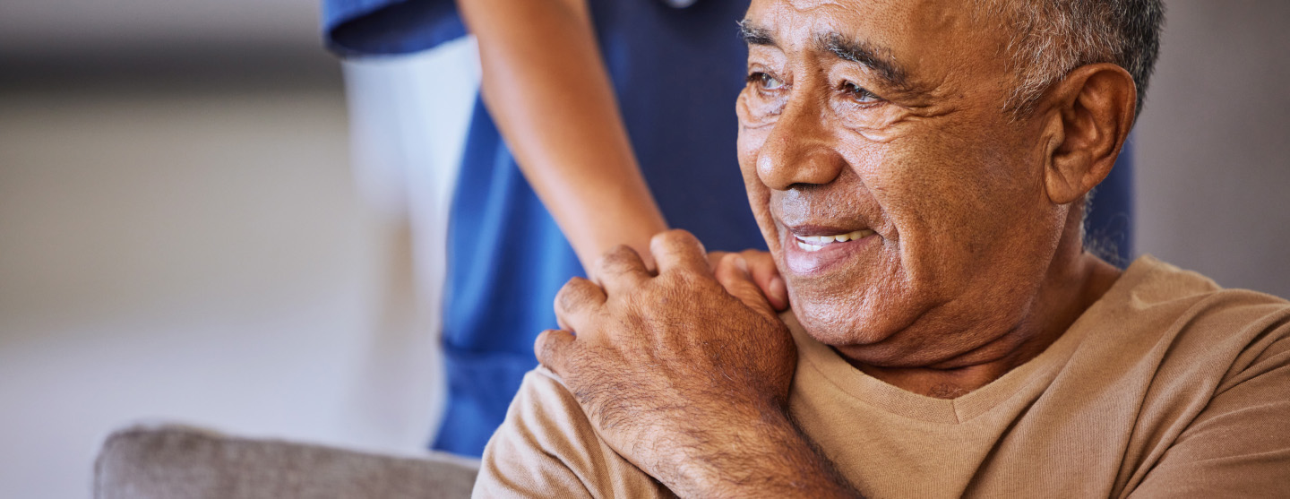 A senior man smiles and gets support through the touch of his shoulder from a healthcare provider.