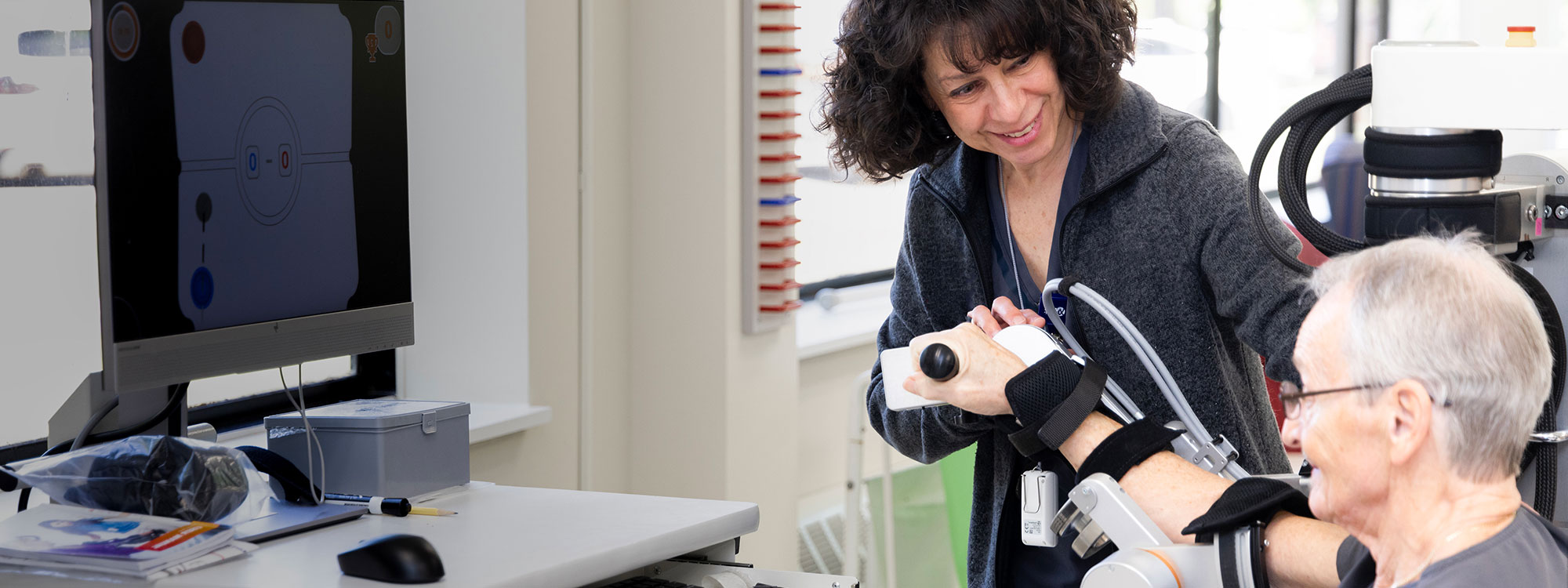 A healthcare provider smiles while helping a patient use an adaptive technology on their arm in a physical therapy setting