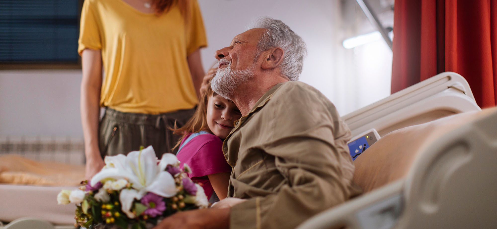 Young girl giving her grandpa flowers while visiting him in hospital with her mother